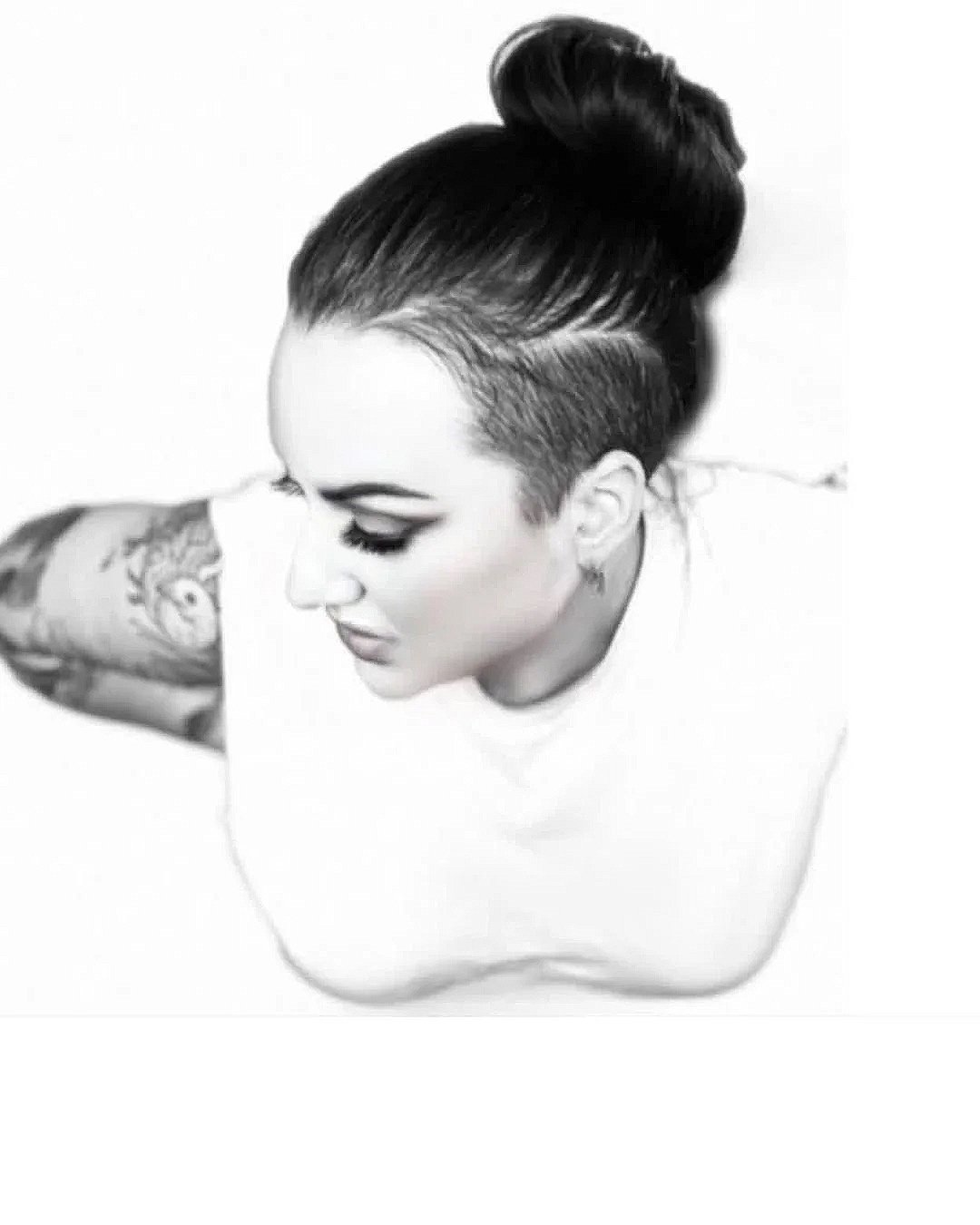 Black and white photo of a woman with her hair styled in a high bun and shaved sides, tattoos on her arm, and wearing a white t-shirt.