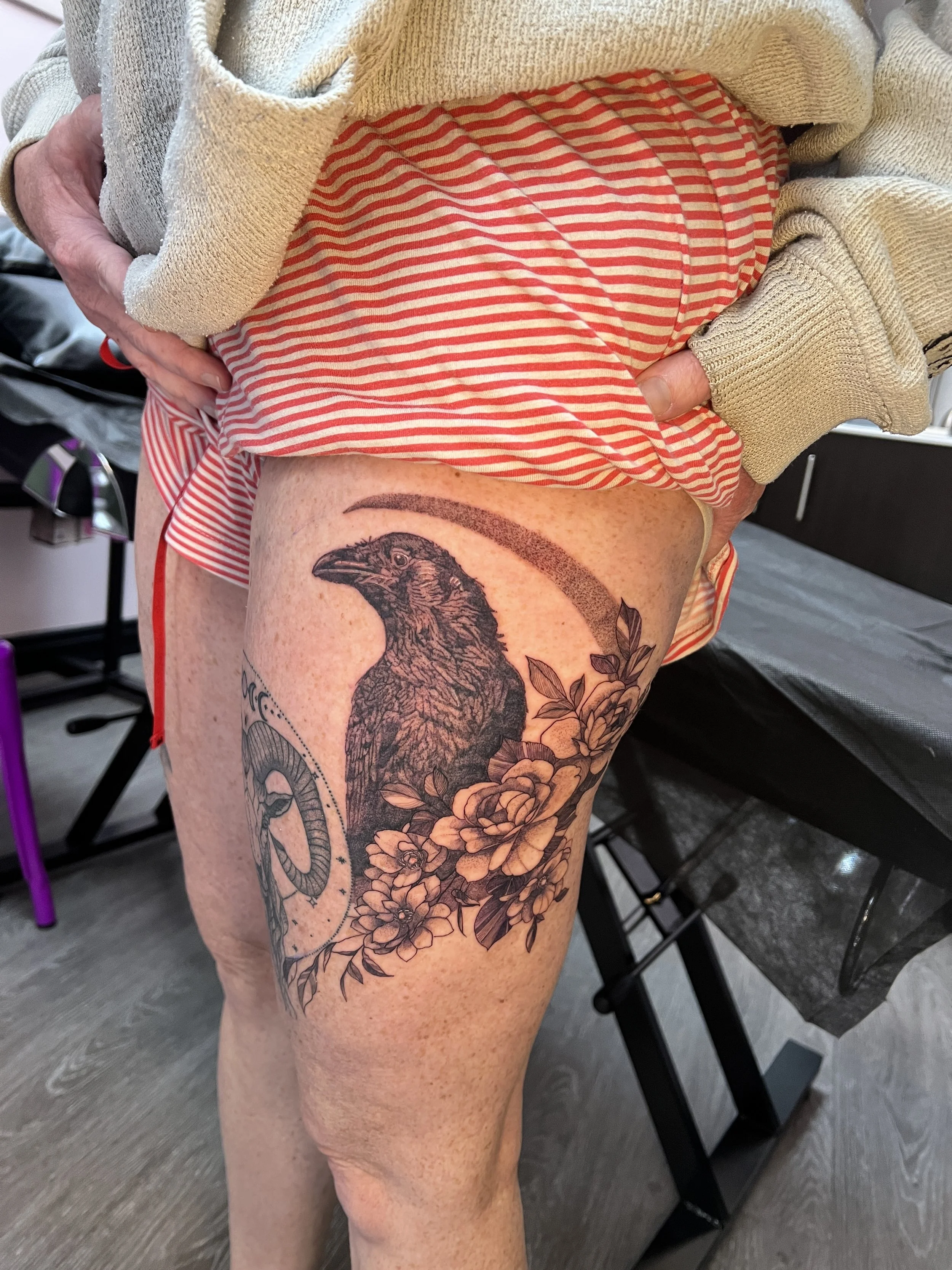 A person's upper thigh showing a detailed black and gray tattoo of a raven's head with flowers around it.