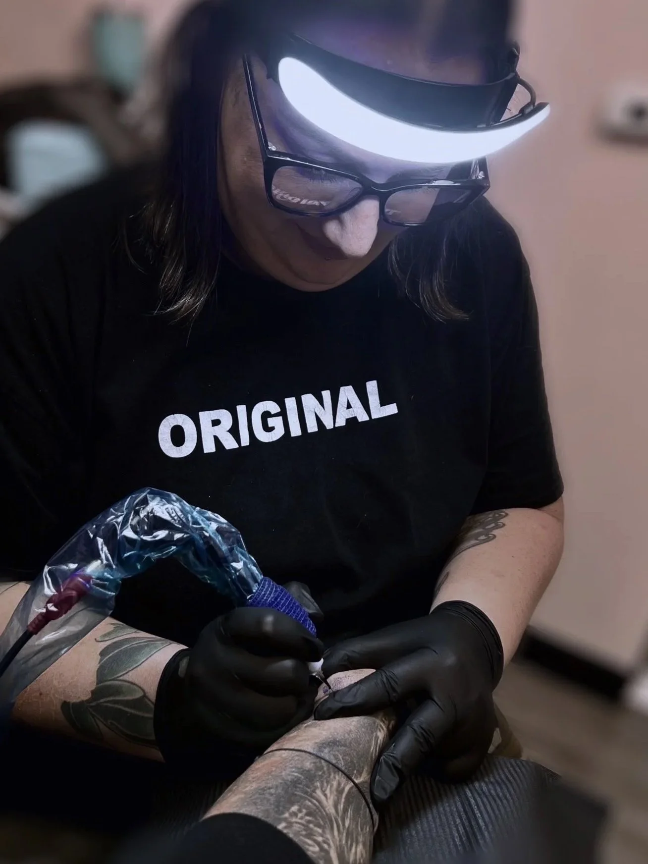 A person wearing glasses and a mask tattooing another person's tattoo on their leg, with a focused expression and tattoo equipment visible.