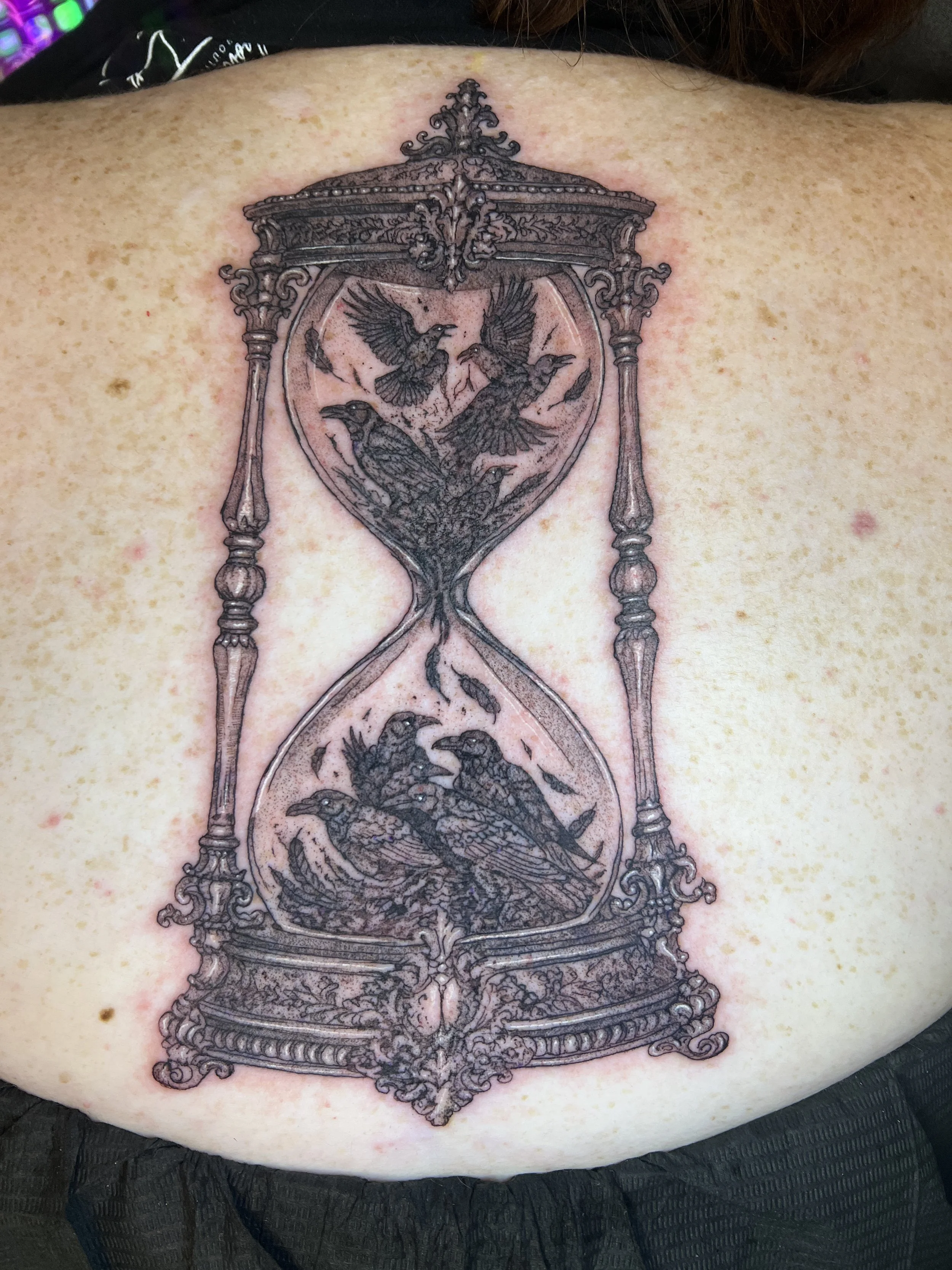 A tattoo of an ornate, vintage hourglass with detailed birds and decorative elements, located on the person's back.