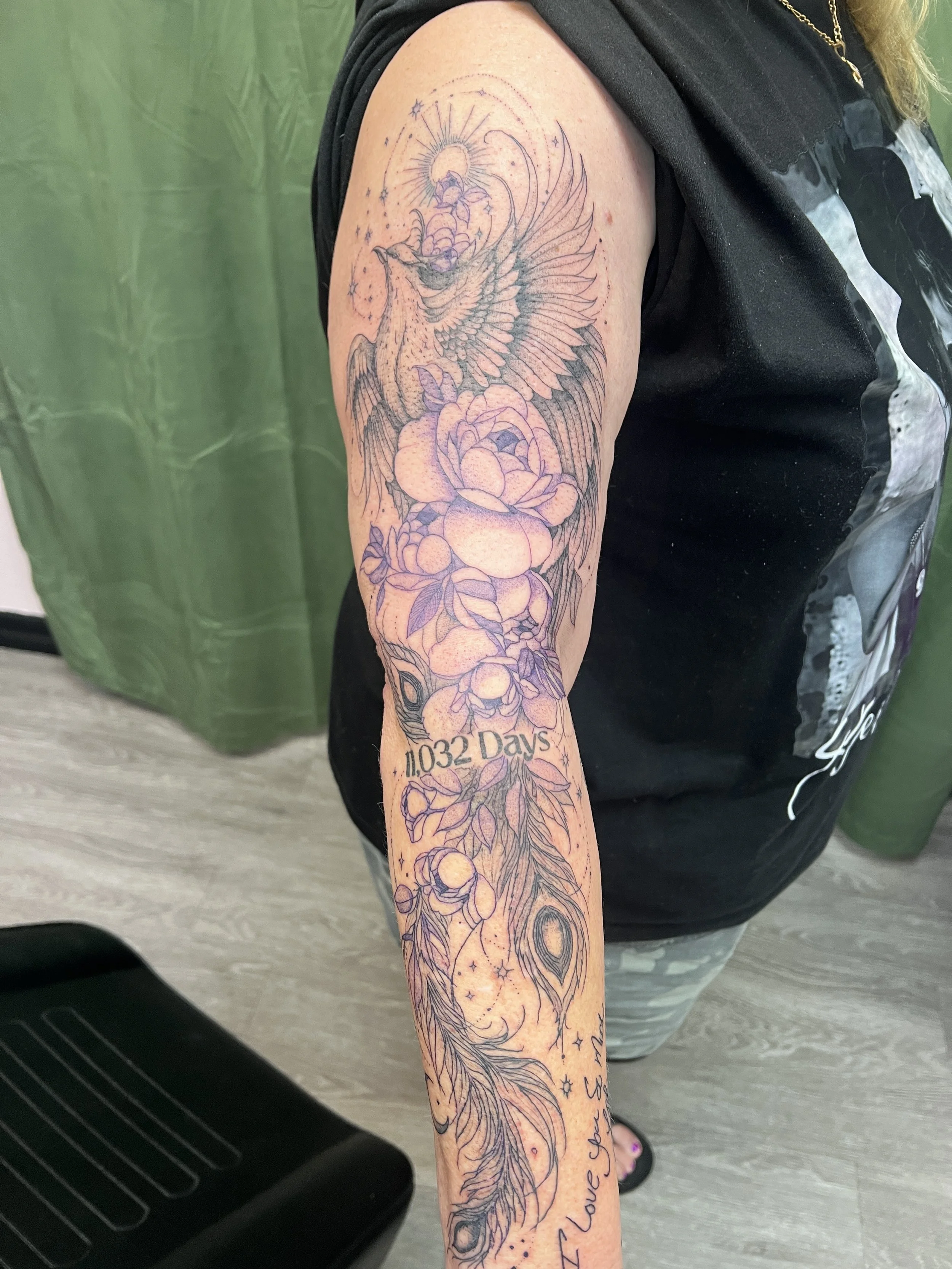 Tattoo of a phoenix rising above pink and purple flowers on a person's upper arm.