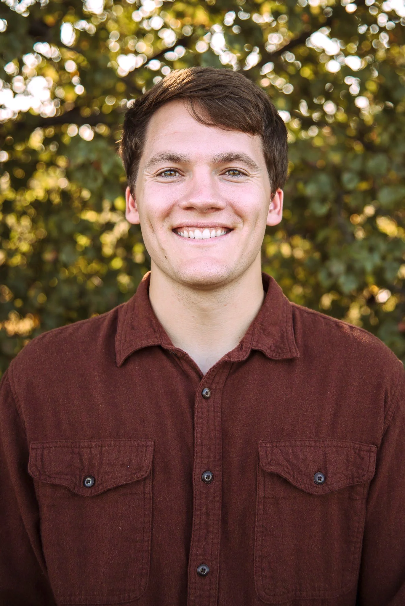 Young man with short brown hair smiling outdoors with blurred trees in the background.