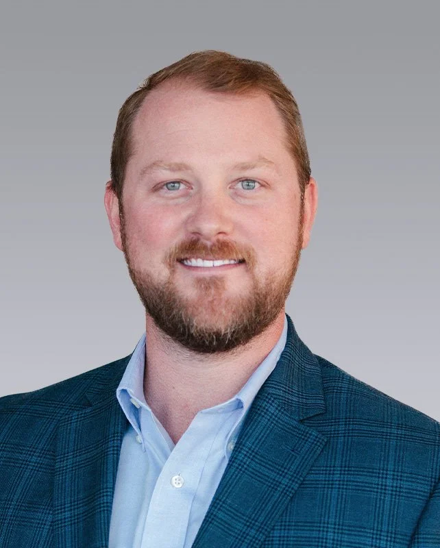 A professional headshot of a man with light brown hair and a beard, wearing a blue plaid blazer and a light blue collared shirt, smiling against a plain gray background.