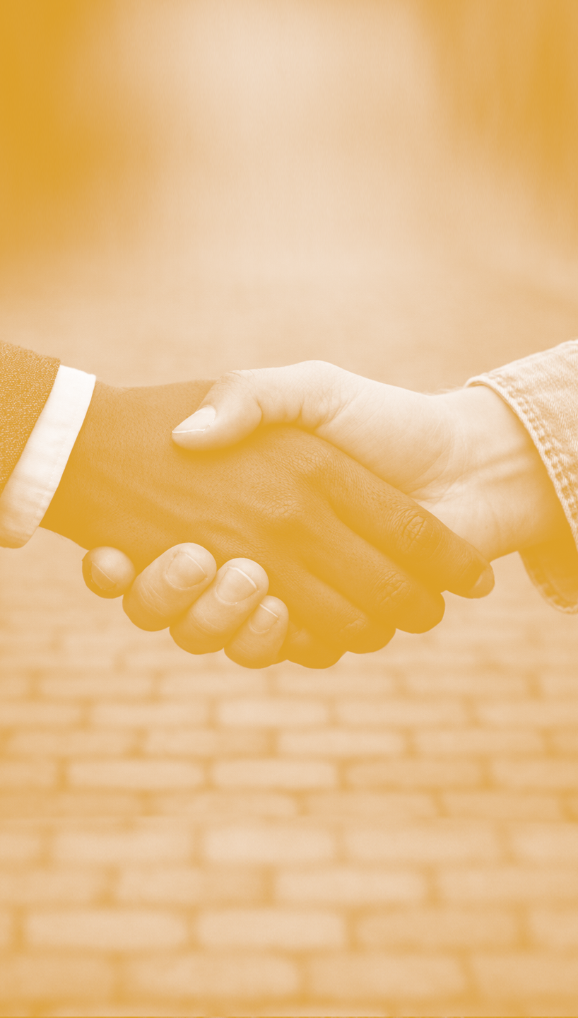 Two people shaking hands, one wearing a suit with a white shirt and the other in a casual denim shirt, on a brick floor background.