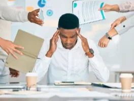 A man sitting at a desk with hands on his head, surrounded by multiple hands passing documents and a laptop, indicating a stressful work situation.