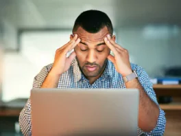 Man sitting at a desk with hands on his forehead, looking stressed in front of a laptop