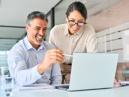 Two people, a man and a woman, smiling and working together on a laptop in an office setting.