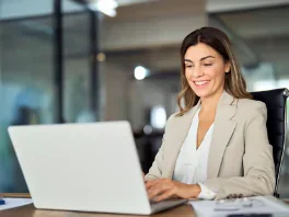 A woman sitting at a desk, smiling, using a laptop in an office setting.