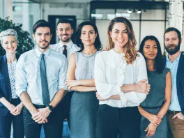 A diverse group of seven professionals standing together in an office environment, smiling at the camera.
