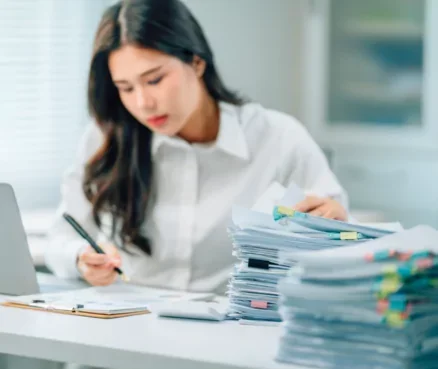 Woman in white shirt working at desk with large stacks of papers and files