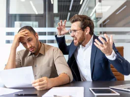 Two men sitting at a table in a modern office, engaged in a heated argument or intense discussion. One man looks frustrated while the other appears angry and is gesturing aggressively.