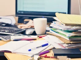 Cluttered desk with many papers, notebooks, and writing utensils in front of a computer monitor.