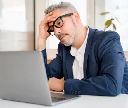 A man with gray hair and glasses, dressed in a blue blazer and white shirt, sitting at a desk with a laptop, appearing stressed or focused.