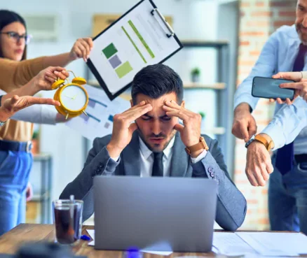 Man sitting at a desk, appearing stressed, surrounded by people holding office items in a busy office setting.