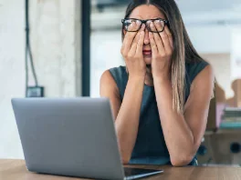A woman sitting at a desk with a laptop, holding her face with both hands and looking surprised or overwhelmed.
