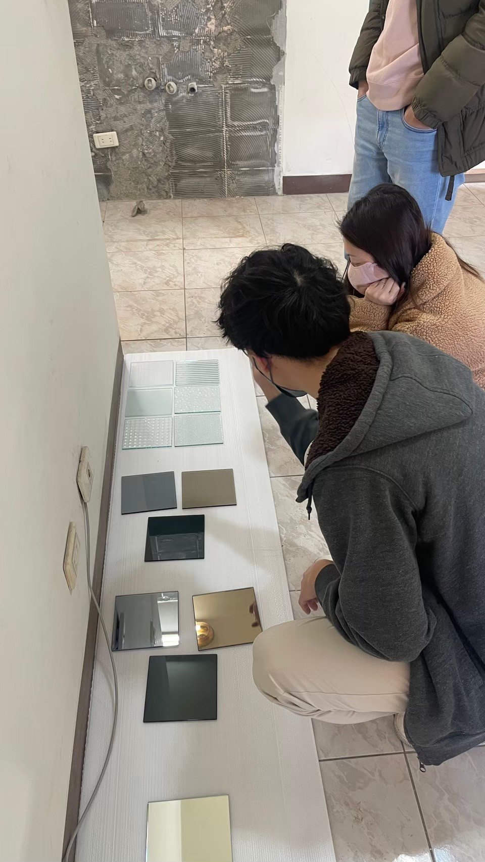 People examining tile samples on the floor near a partially torn-down wall in a room under renovation.