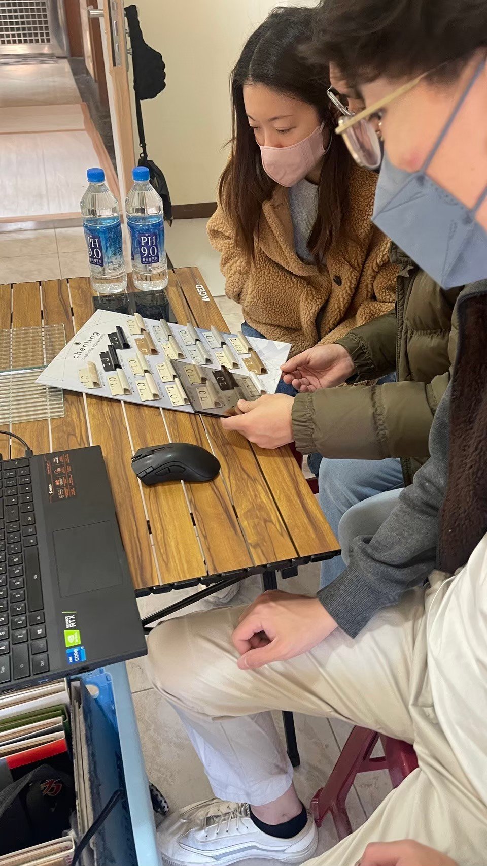 People sitting around a wooden table with a laptop, examining color samples and water bottles.