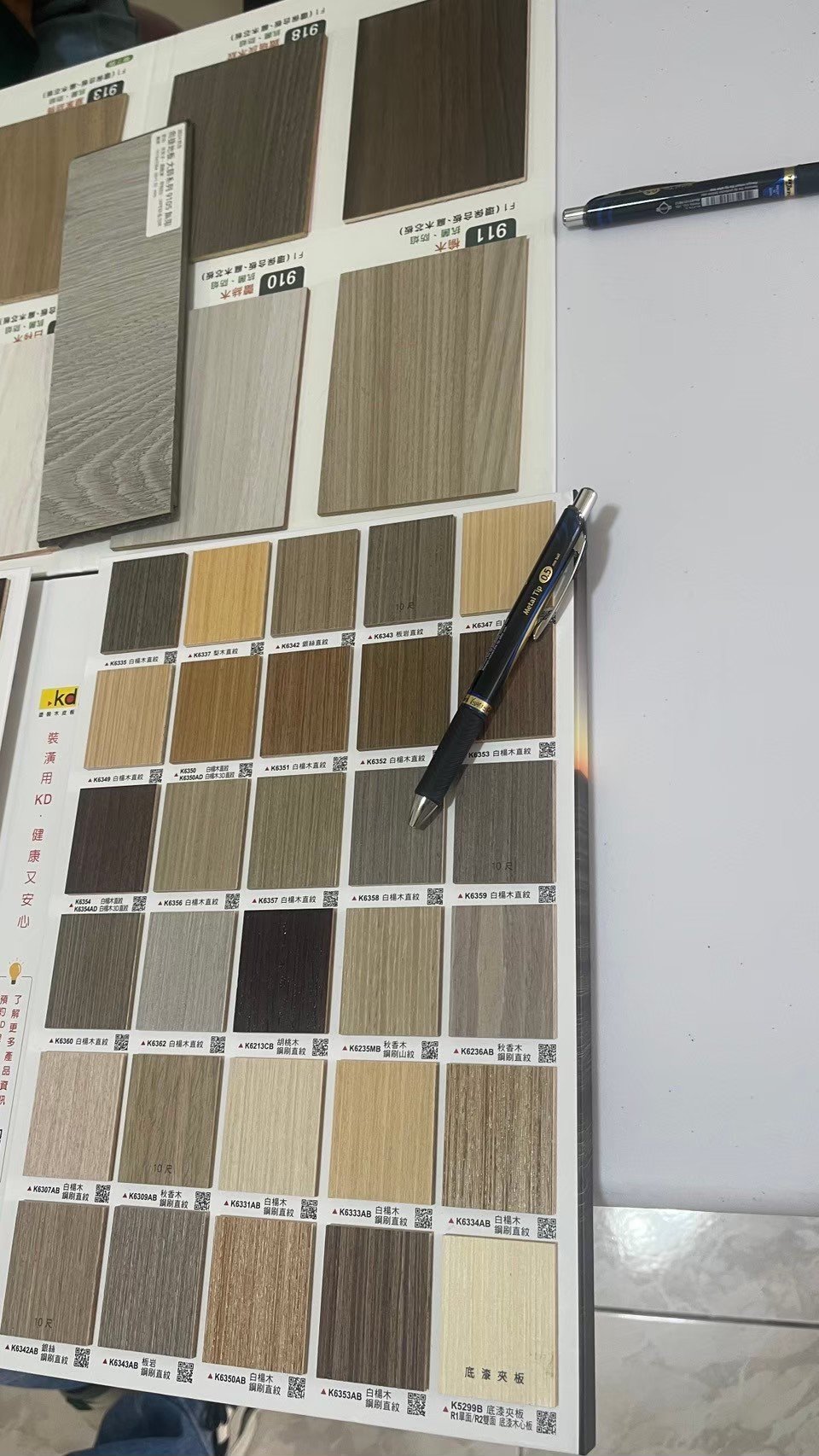 A collection of wooden laminate samples displayed in a grid with various shades and textures. Two black pens are placed on the sample booklet for reference.