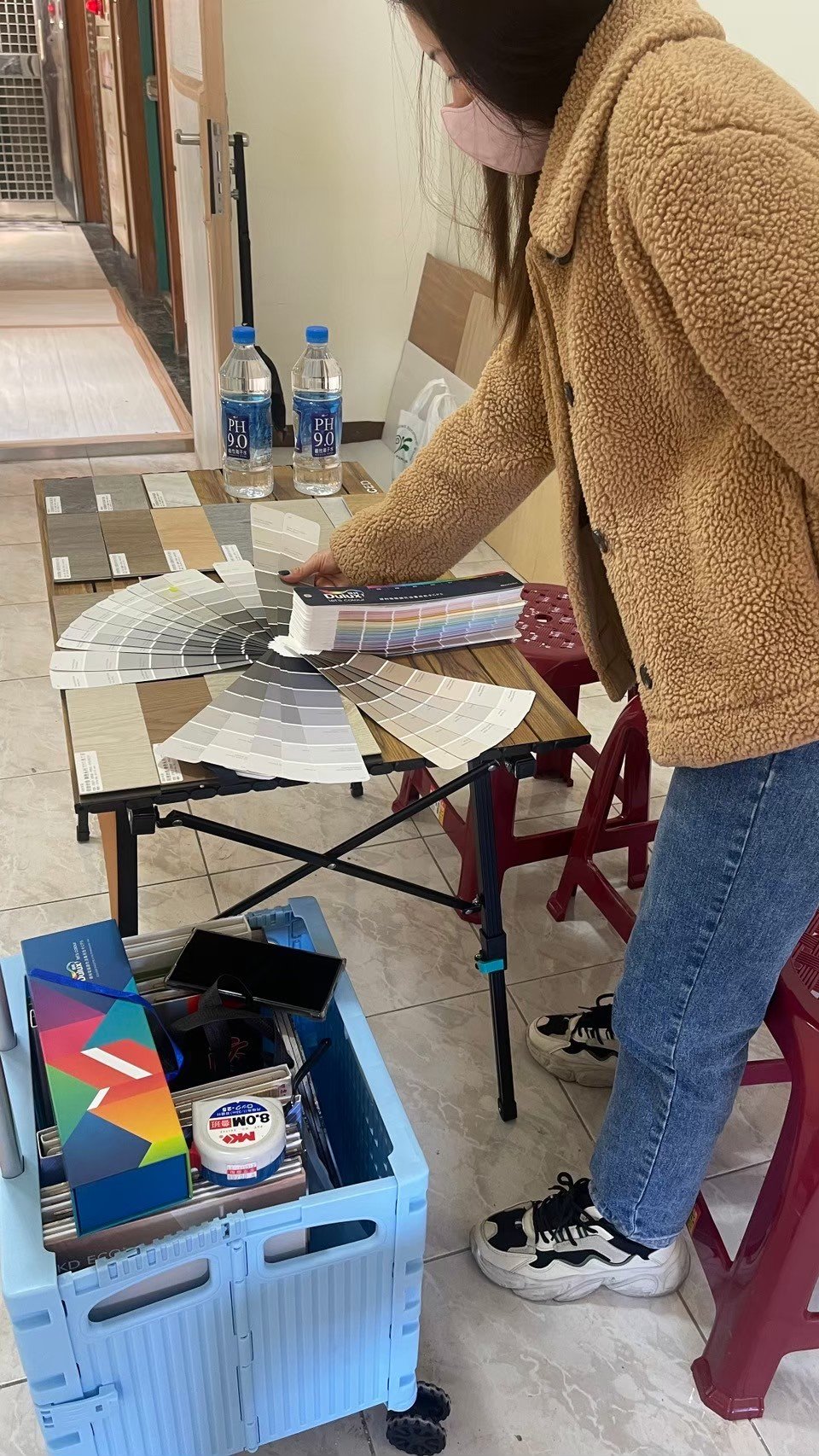 Person selecting colors from a palette fan on a wooden table with water bottles and a storage box nearby.