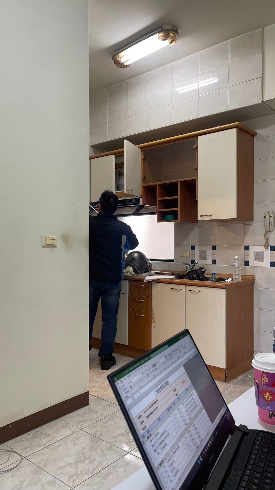Person in a kitchen setting working with open cabinets, a laptop with a spreadsheet open in the foreground, and kitchen items like a helmet and cup nearby.