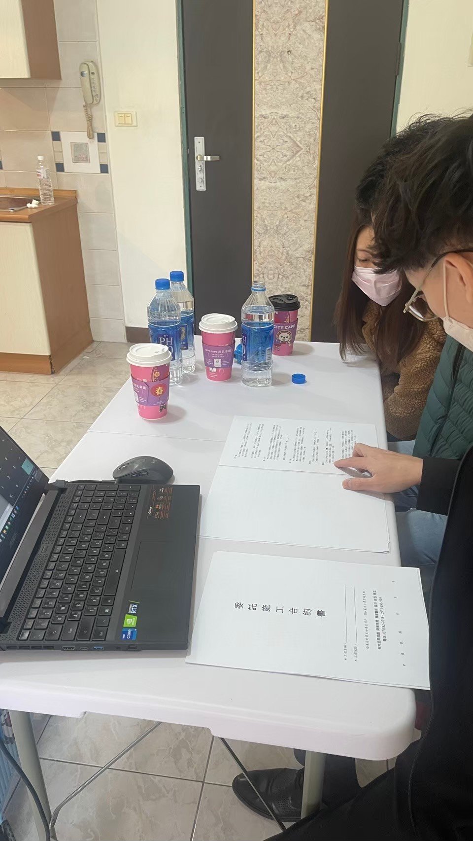 Two people at a table with documents, a laptop, bottled water, and coffee cups. The people are wearing masks and reviewing the papers.