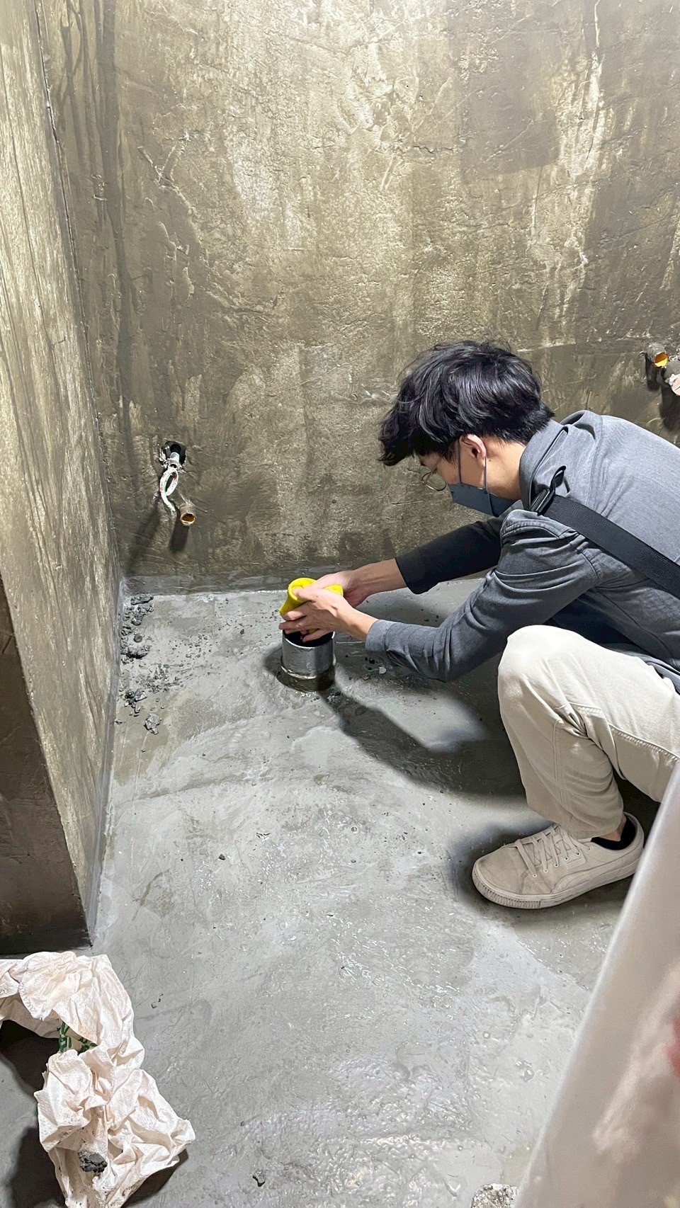 Person crouching on concrete floor, working on a plumbing fixture in a dimly lit, unfinished room.