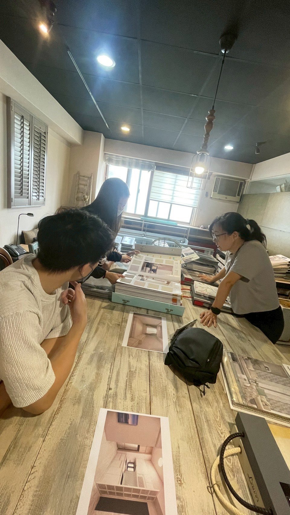 Three people reviewing home design blueprints and materials at a wooden table, with printed designs and samples spread out, in a modern office setting.