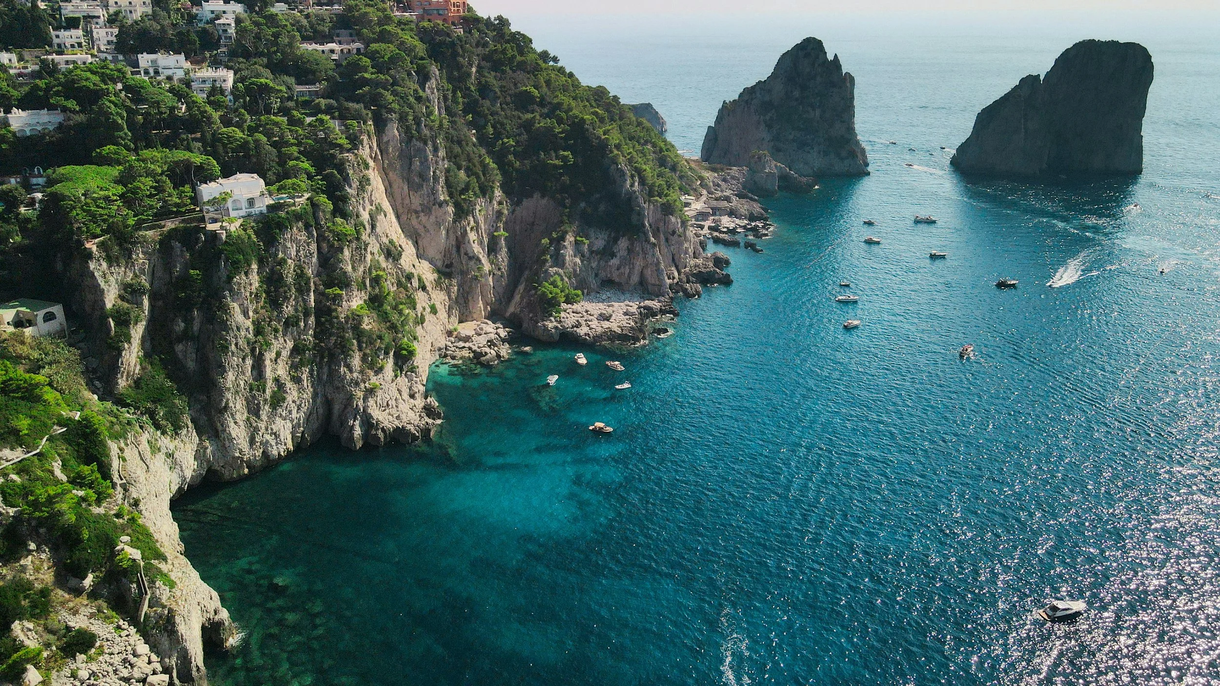 Aerial view of a coastal landscape with steep cliffs covered in lush green vegetation, white buildings on the hillside, and a deep blue sea with boats scattered across the water near two large rock formations