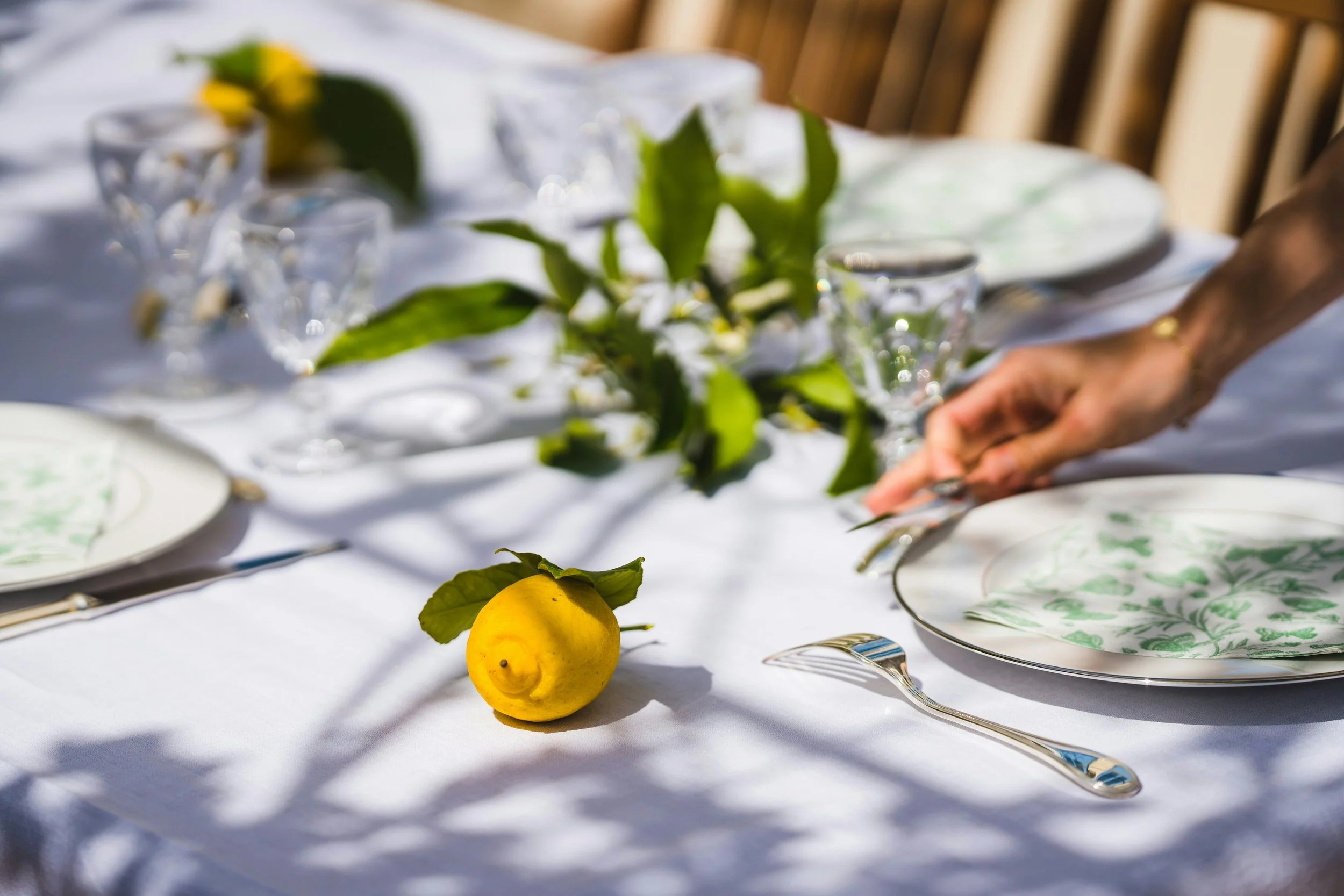 A beautifully set outdoor dining table with white tablecloth, green patterned plates, silverware, clear glassware, a yellow lemon with green leaves, and green foliage in the center, with a person's hand reaching for a plate.
