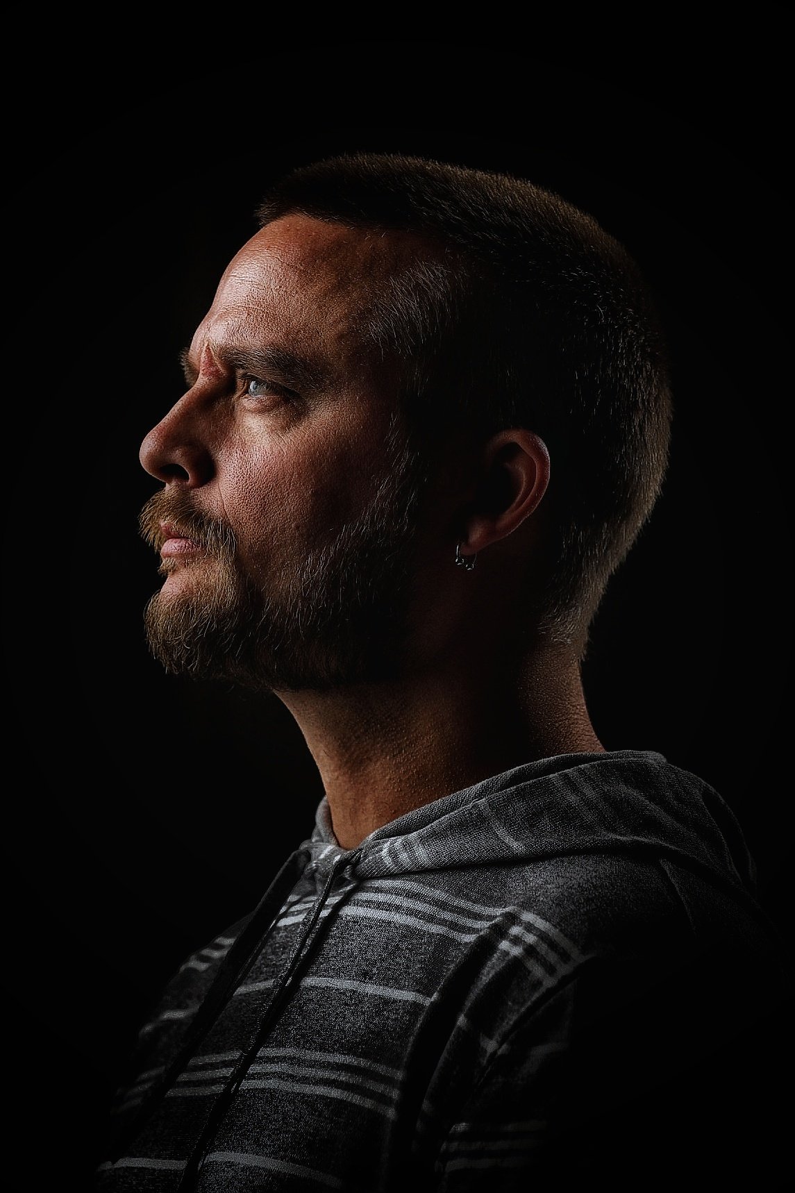 Profile of a man in Chattanooga with short dark hair, beard, and earring, wearing a gray plaid hoodie against a black background for a headshot session.