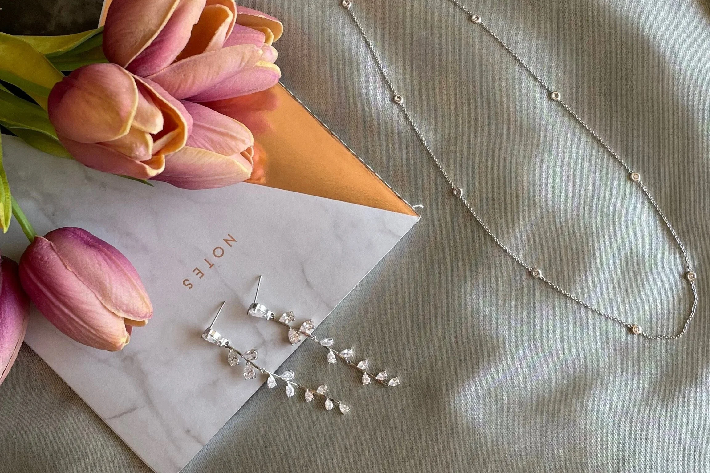 Curated selection of diamond jewellery with notes and flowers on grey linen