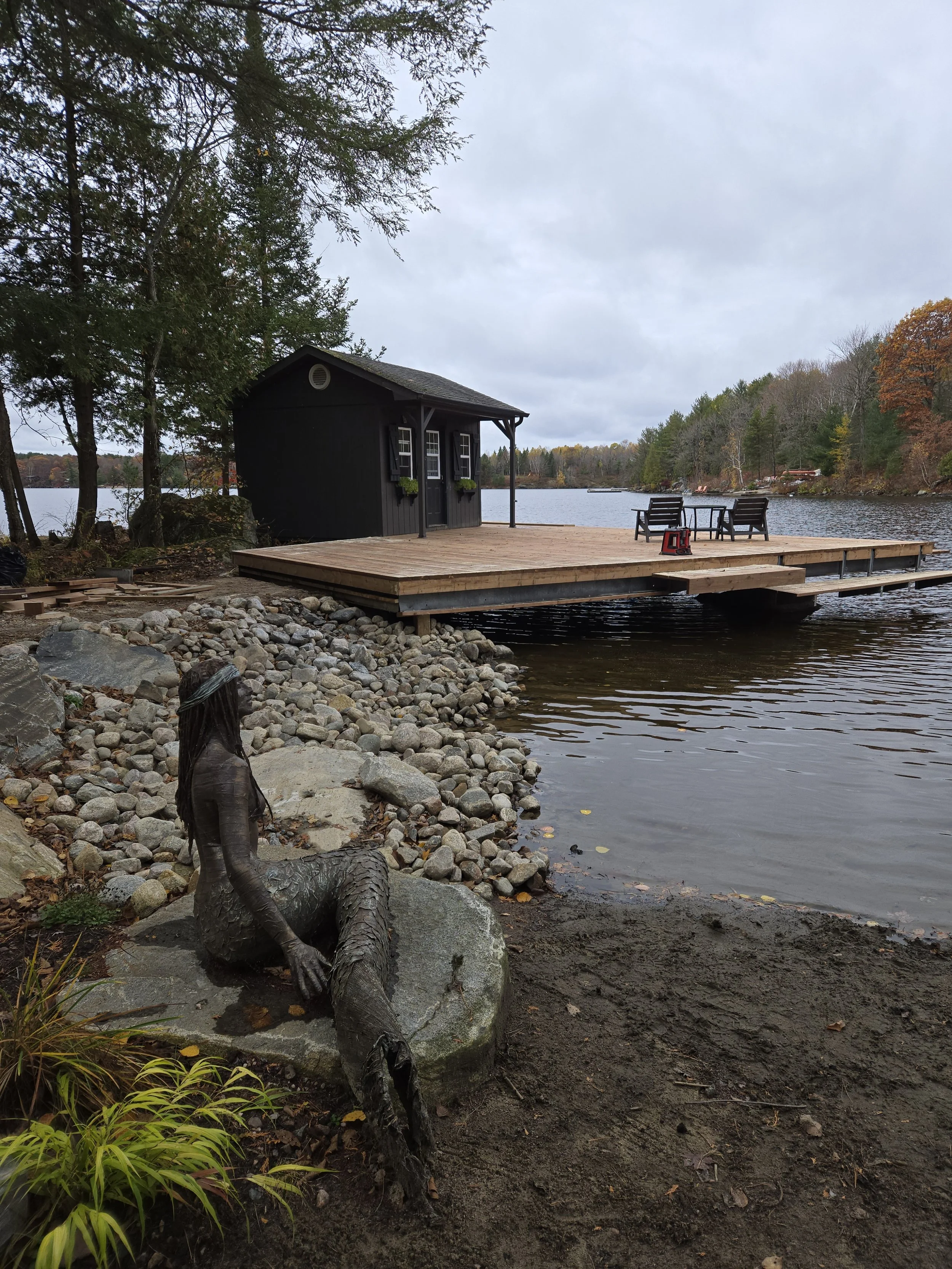 A lakeside scene featuring a small black cabin on a wooden deck, with outdoor seating and a small red stool. A decorative mermaid sculpture is on a rock near the shore, surrounded by rocks and trees with fall foliage, with a cloudy sky overhead.