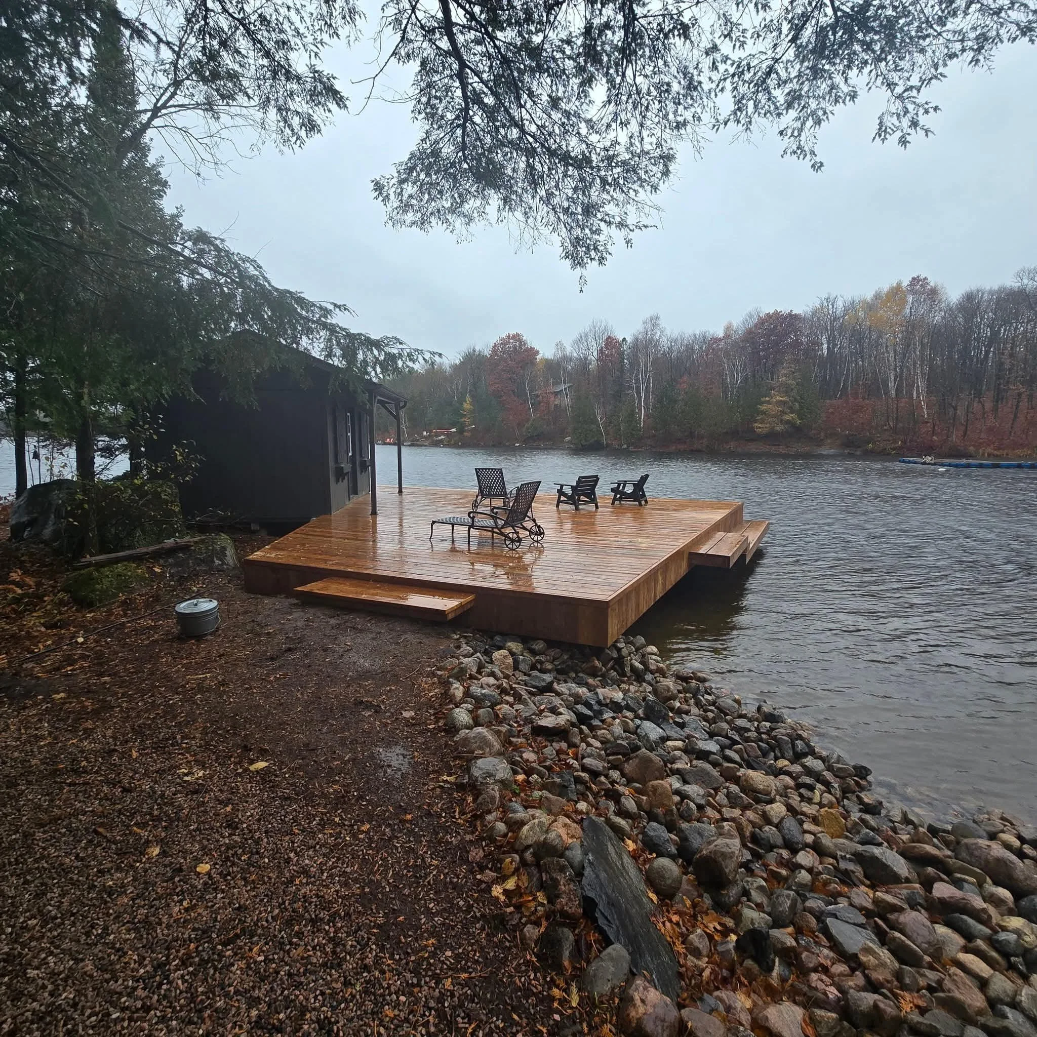 Rainy day at a lakeside with a wooden deck extending over the water, surrounded by rocks and trees, with outdoor chairs and a small black shed on the deck.