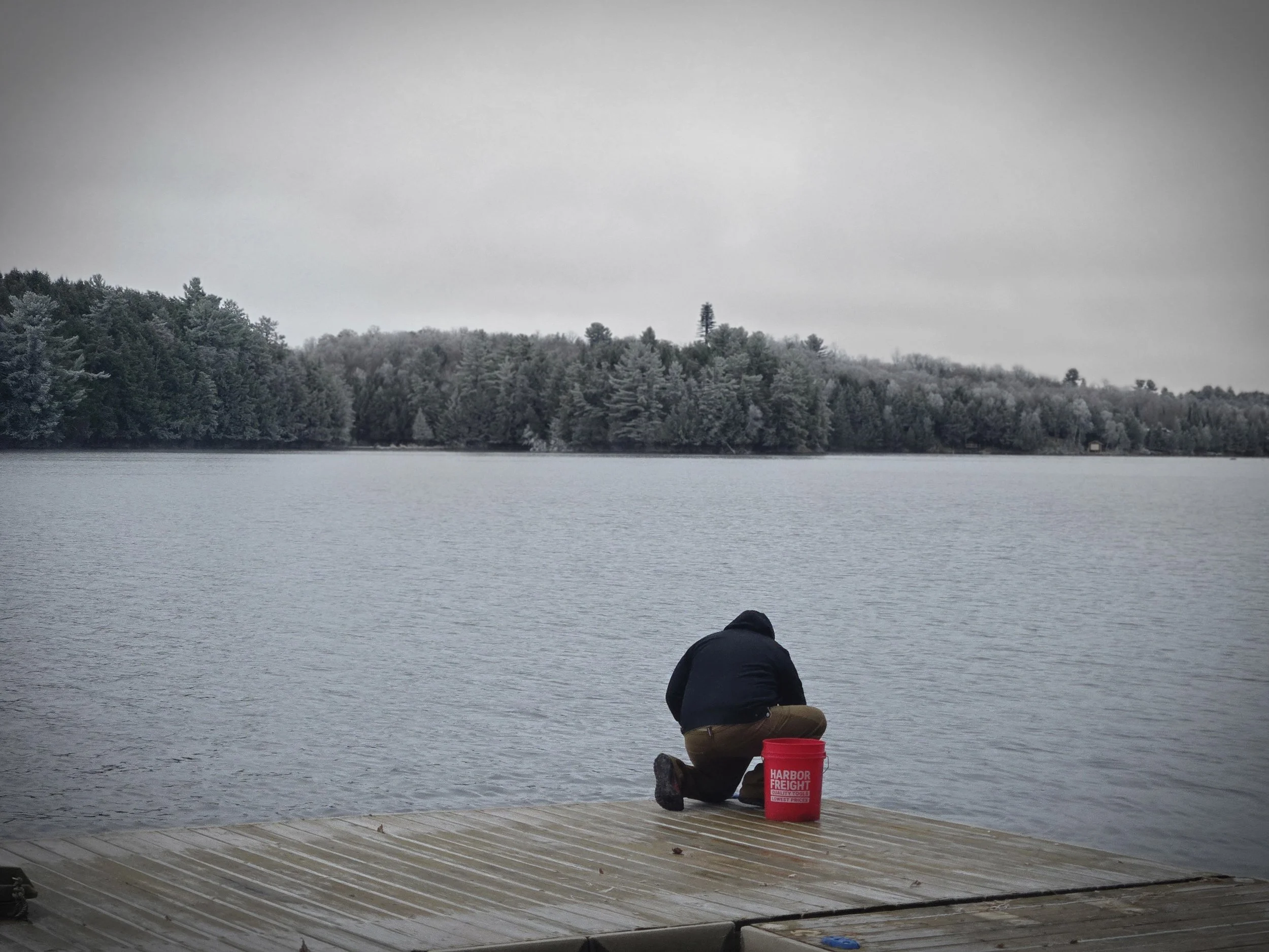A person kneeling on a wooden dock near a body of water with a red bucket labeled 'Harbor Freight' beside them, and a forested shoreline under an overcast sky in the distance.