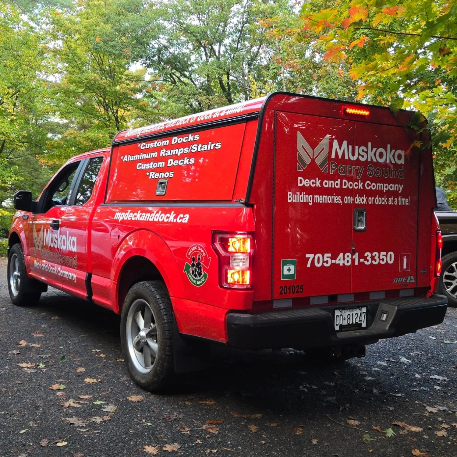 Red company truck for Muskoka Parry Sound Deck and Dock Company parked outdoors on a paved area with fallen leaves, surrounded by trees with green and some orange leaves, displaying company information and contact details.
