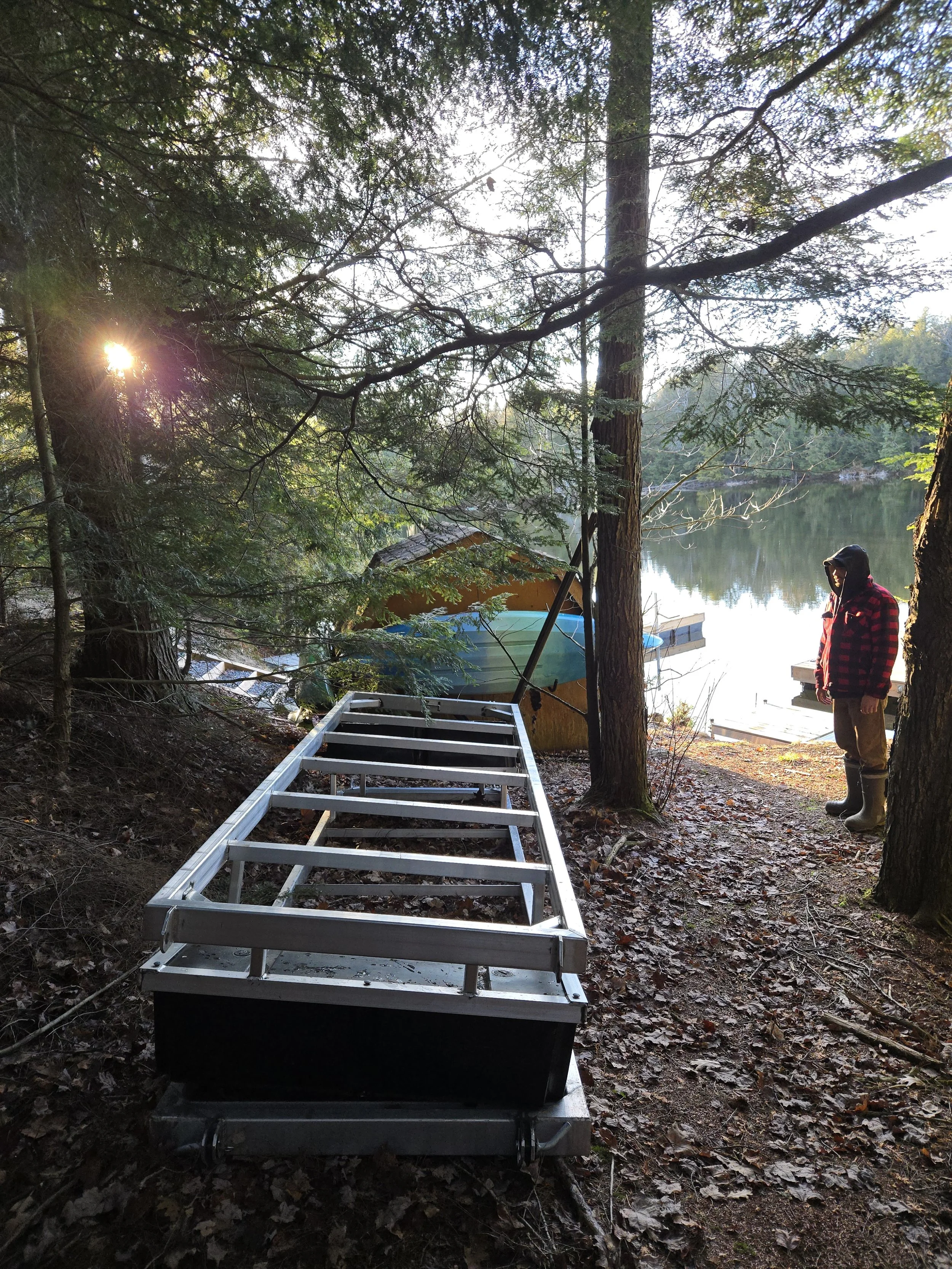 A metal dock frame lying on the ground in a wooded area near a lake, with a person wearing a red and black checkered jacket and boots standing nearby, looking towards the water.