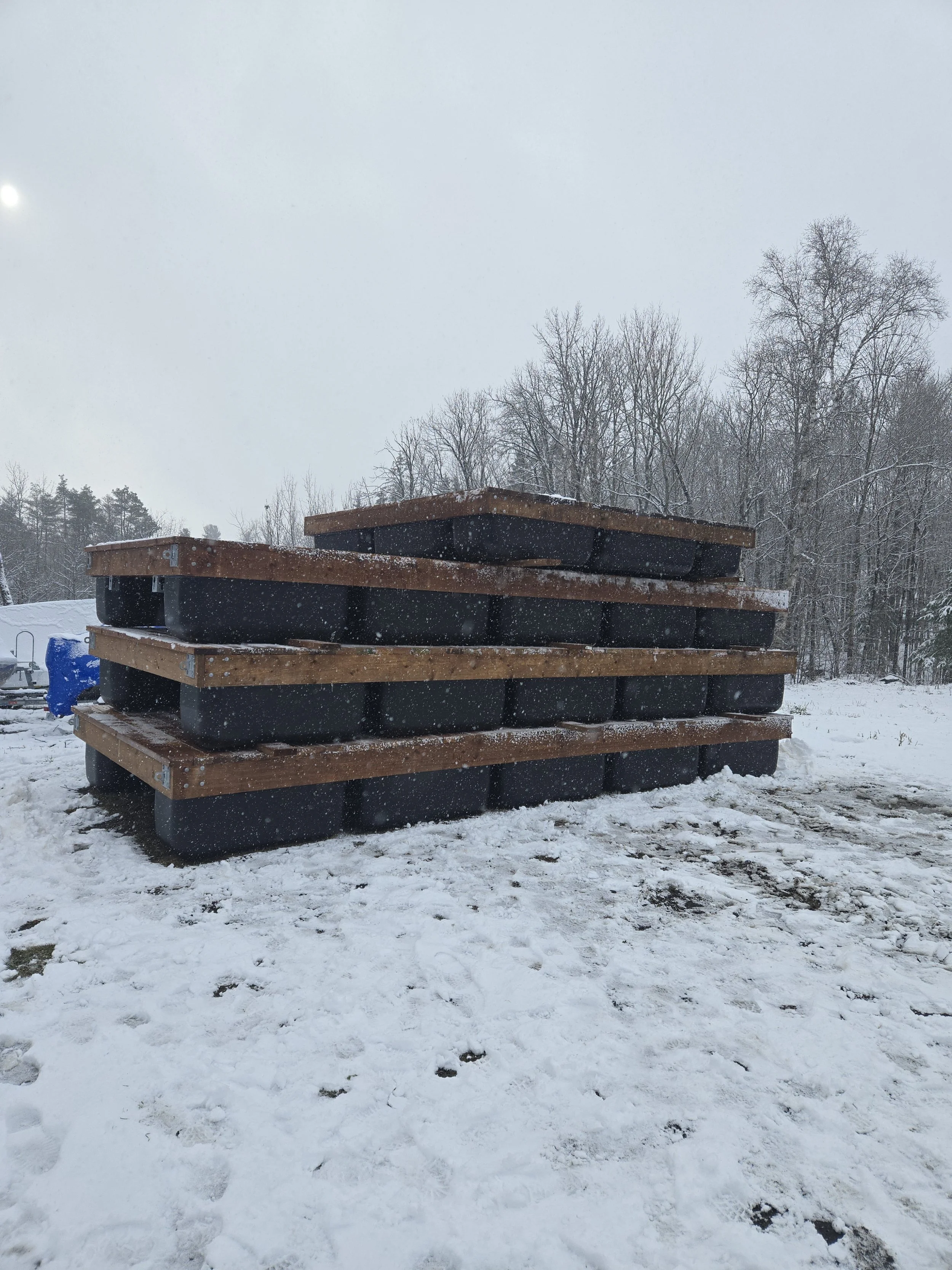 Stacked black plastic containers with wooden frames outside in snowy weather, with leafless trees in the background.