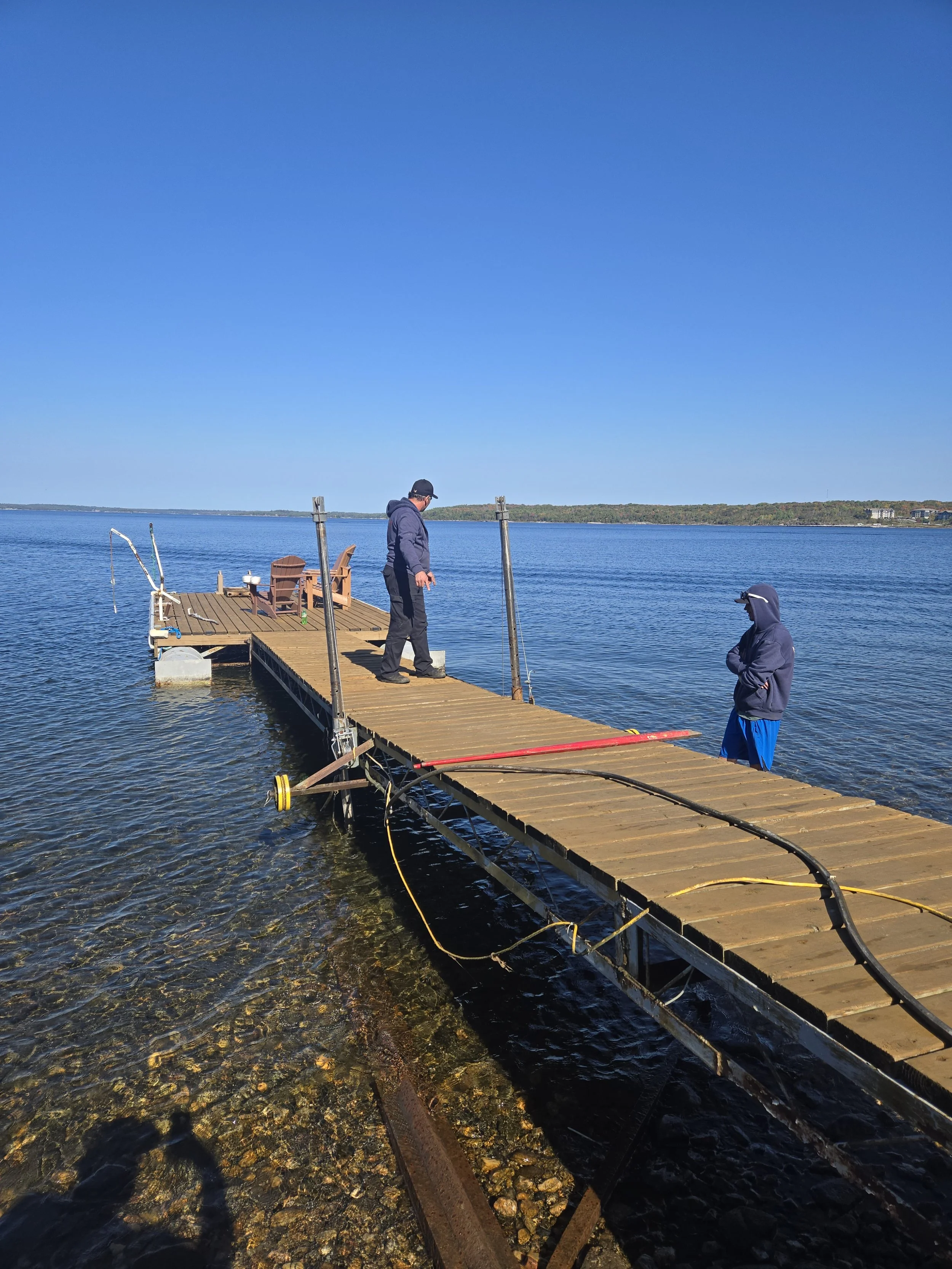 Two men standing on a wooden dock extending into a body of water on a clear, sunny day, with chairs and a bench at the end of the dock.