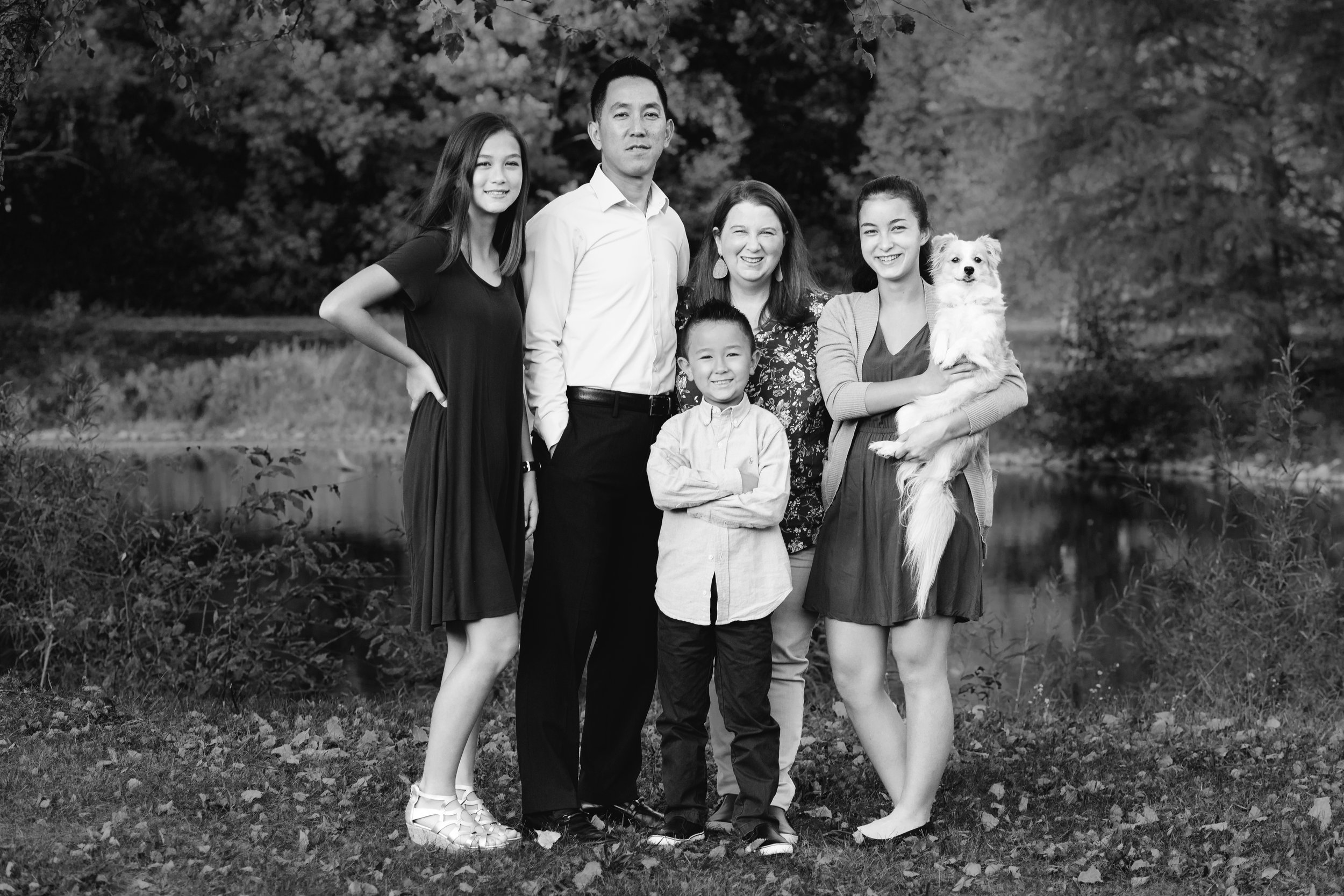 Black and white photo of a family of six people, with three girls, one boy, and two adults, standing outdoors near a pond and trees. One girl is holding a small white dog.
