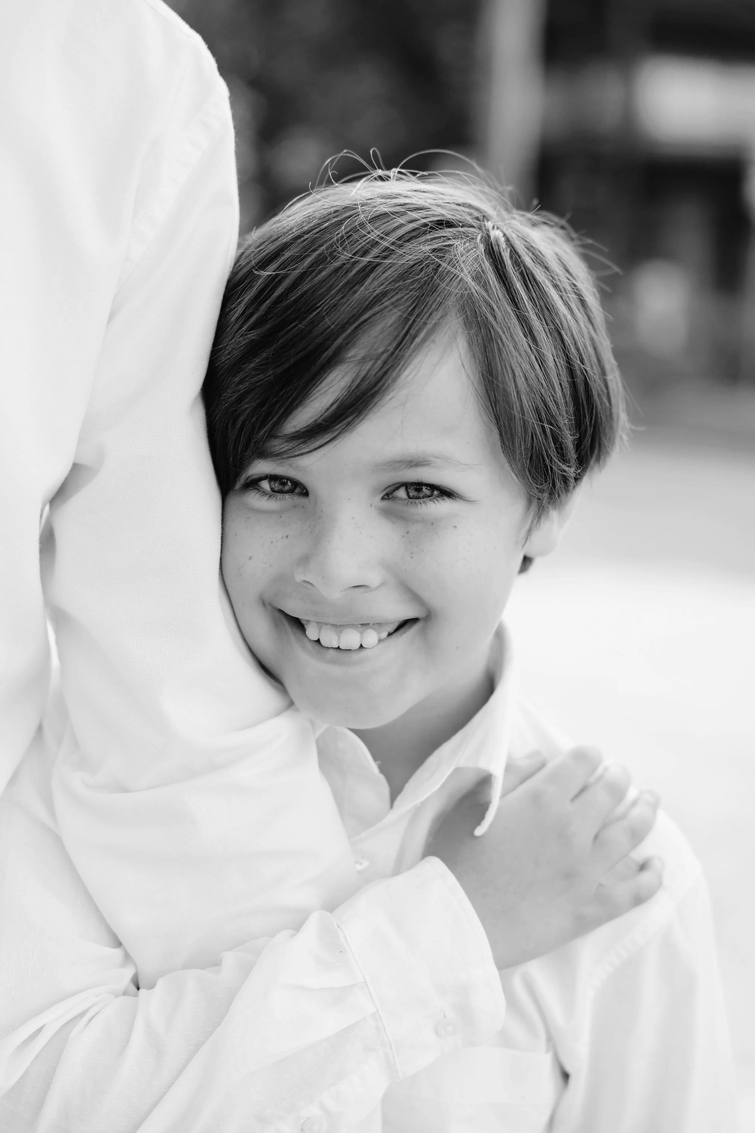 Black and white close-up of a smiling young boy with light-colored hair, wearing a white shirt, being embraced by an adult.