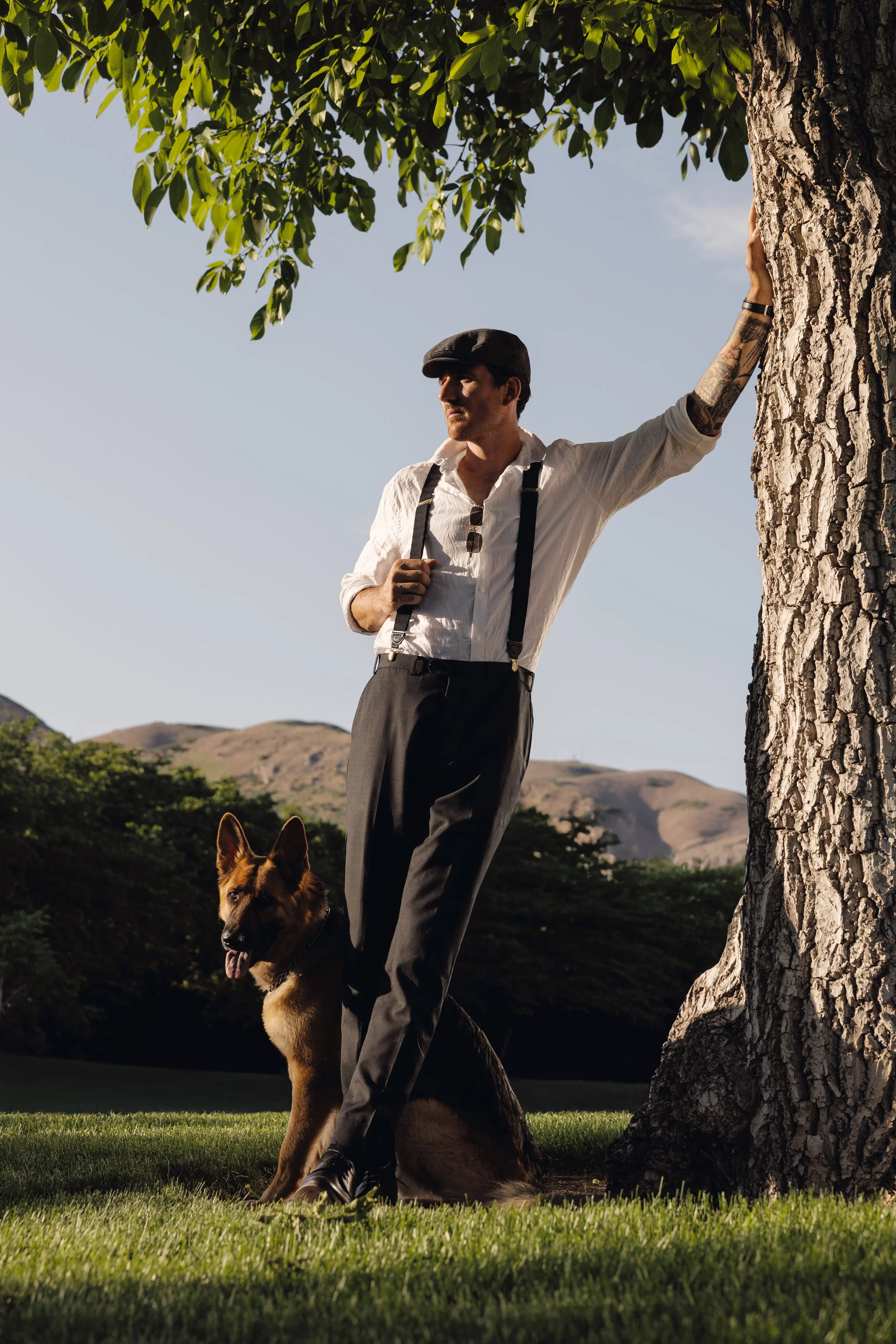 A man with a tattooed arm wearing a white shirt, black suspenders, and a black cap, leaning against a large tree with one hand, standing on the grass with a German Shepherd dog sitting nearby, under a partly cloudy sky with distant hills.