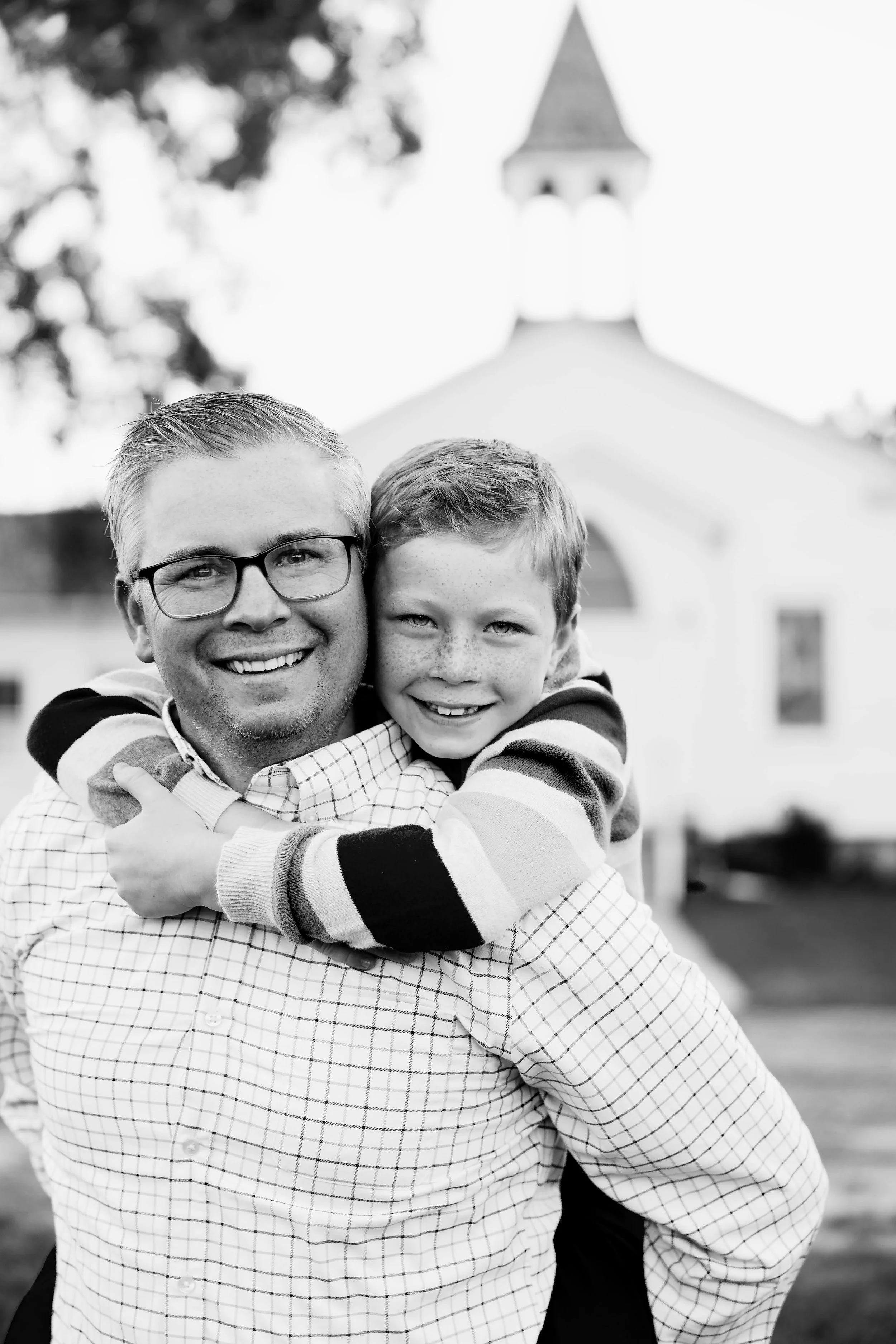 A man and a boy smiling and hugging outdoors, with a church or school in the background.