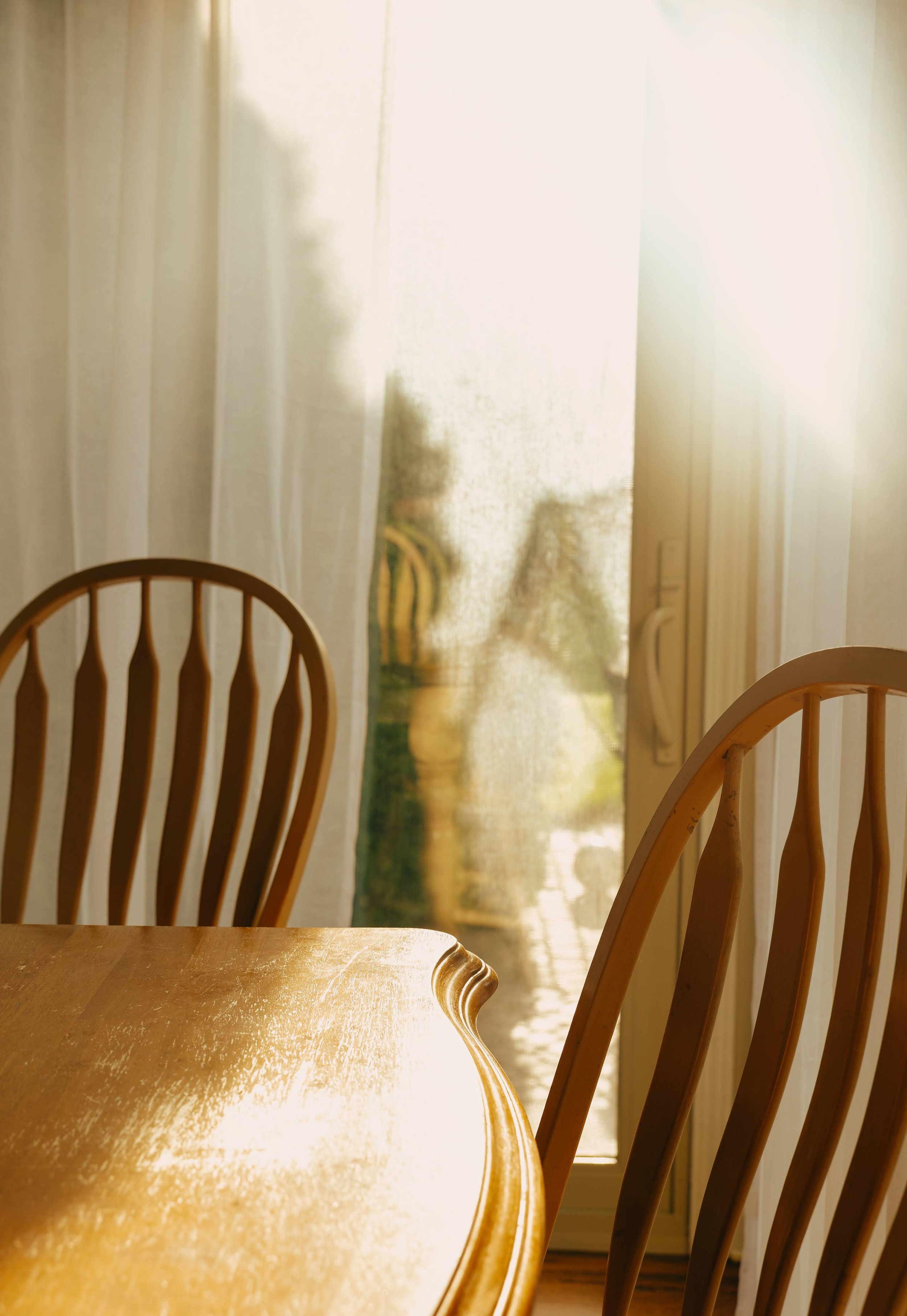 Sunlight streaming through sheer curtains onto a wooden dining table and chairs in a bright dining area.