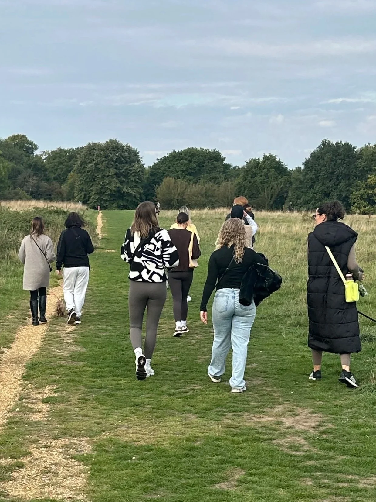 Rivu Club Event Community Walk - Group of people walking on a grassy trail in a open field with trees in the background under a cloudy sky.