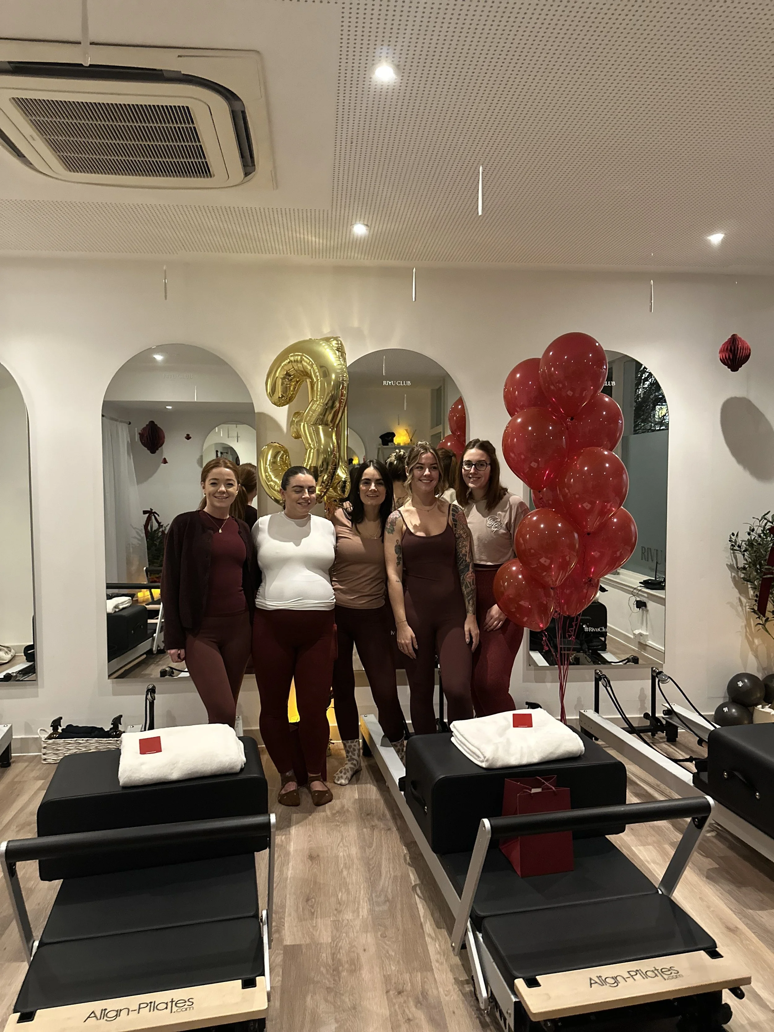 Rivu Club Pilates Party 0 Five women standing together inside a fitness studio, surrounded by red balloons and gold number balloons, while two reformer Pilates machines are in the foreground and mirrors and white walls are in the background.