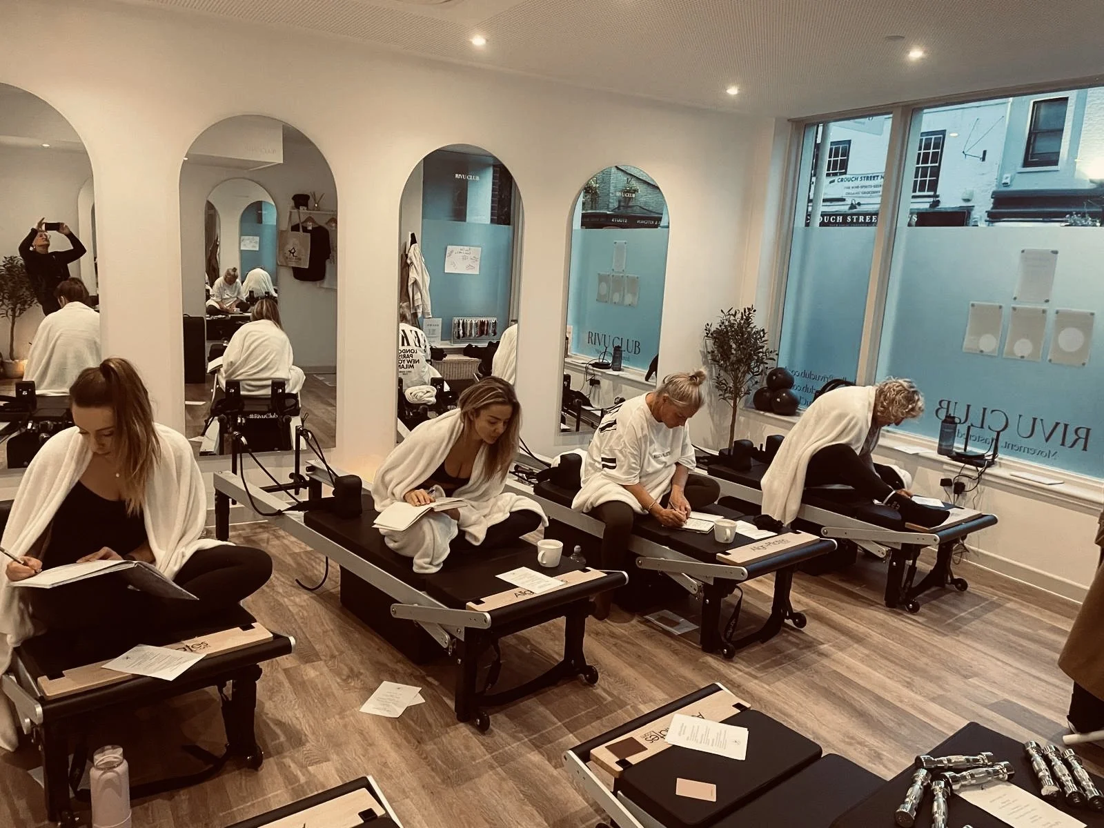 Rivu Club Full Moon PilatesEvent Colchester  - People participating in a Pilates class in a studio, sitting on reformer machines, some reading or writing, with mats, books, and cups nearby, in a well-lit room with large windows and decorative plants.
