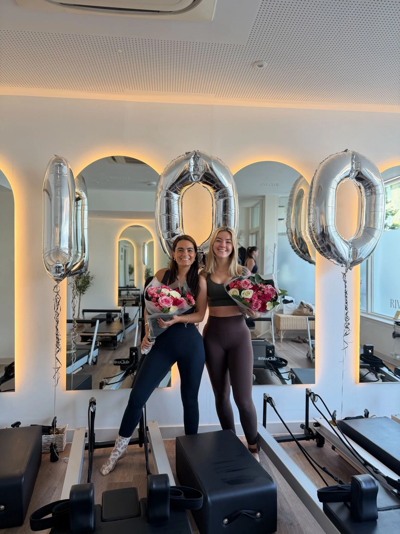 Two women smiling and holding bouquets of flowers standing in front of Pilates reformer machines in a fitness studio decorated with large silver balloons spelling '100' and illuminated mirrors.