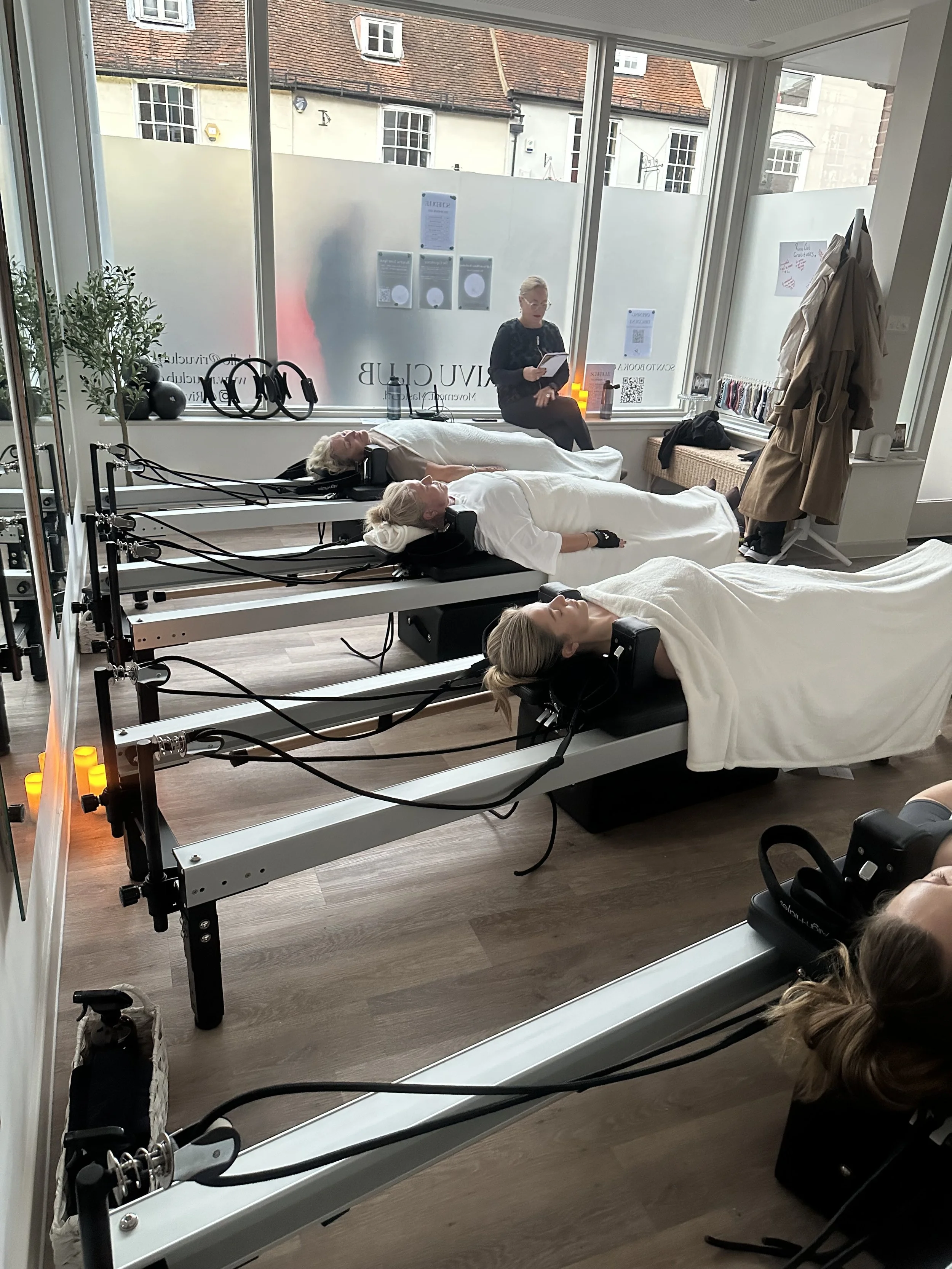 Two women lying on treatment beds with headrests, covered with white sheets, receiving therapy with headgear attachments, in a wellness clinic room. A woman in black listens to music or talks, sitting on a bench next to the window, with a closed-door hanger with beige coat and backpack near her. Windows reveal a view of neighboring buildings.