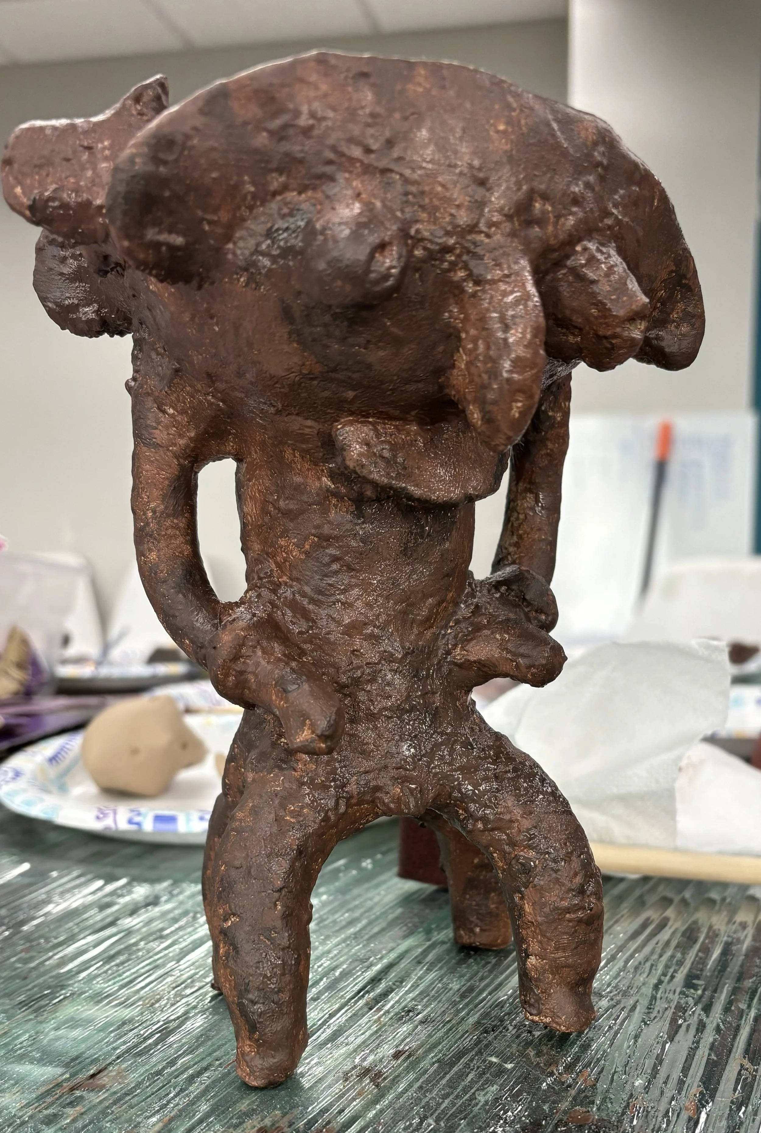 A small figurine sculpture of a humanoid figure with a large head, bent knees, and arms resting on hips, made of a rust-colored material, placed on a table with art supplies and paper towels in the background.