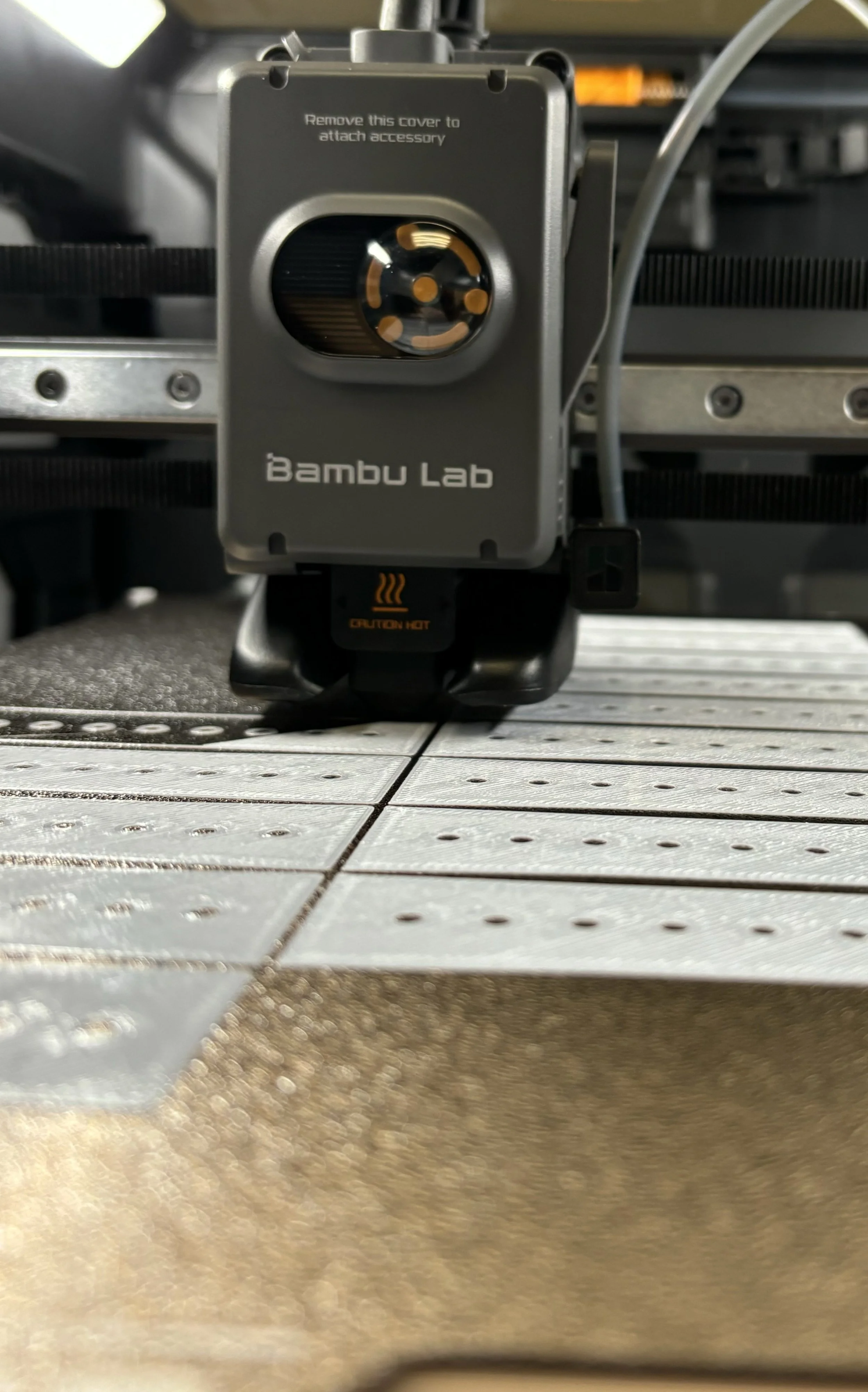 Close-up view of a Bambus Lab 3D printer build platform with a moving print head, which has a warning label 'Caution Hot' and the brand name 'Bambu Lab' on it.